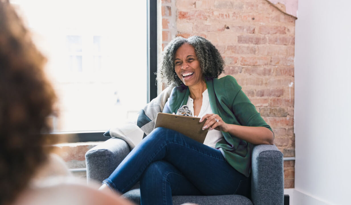 Woman sitting in an armchair, holding a clipboard and laughing. 